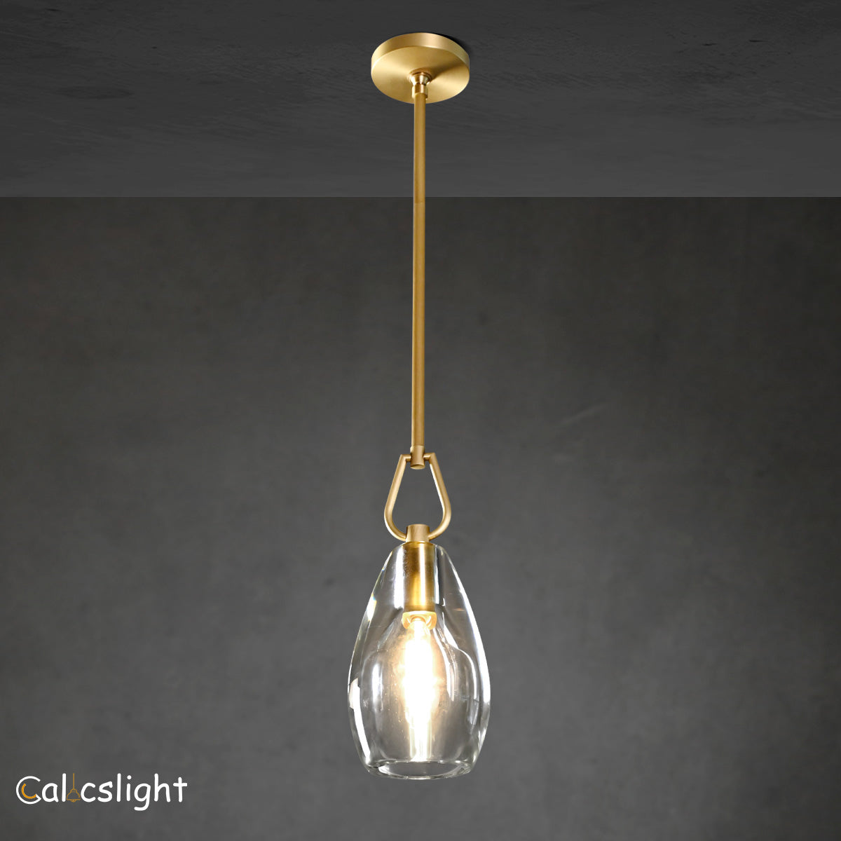 A gold-colored metal pendant light with a clear glass shade, suspended by a chain, against a dark background.
