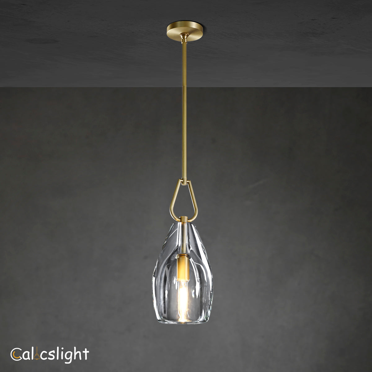 A gold-colored metal pendant light with a clear glass shade, suspended by a chain, against a dark background.
